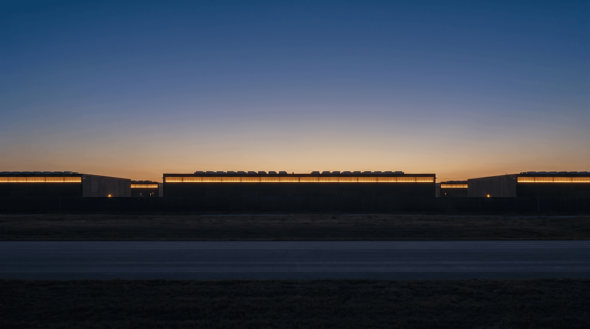 Elevated blue-hour view across the campus roofline — long horizontal bands of charcoal architecture and warm clerestory light