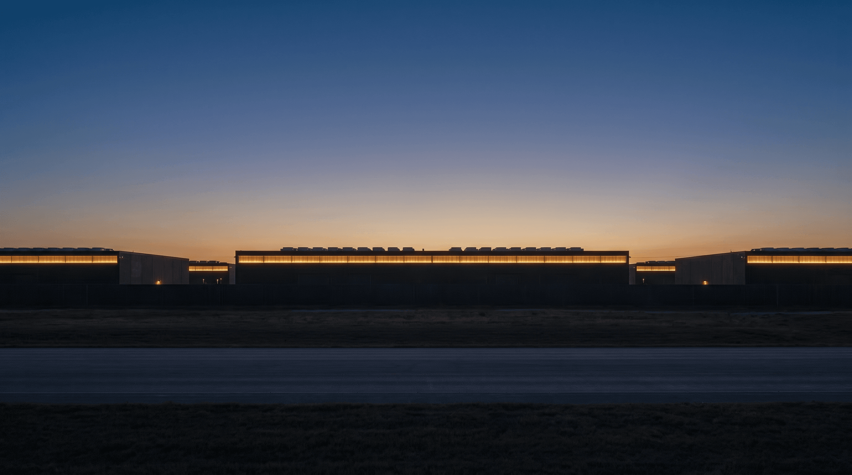 Elevated blue-hour view across the campus roofline — long horizontal bands of charcoal architecture and warm clerestory light