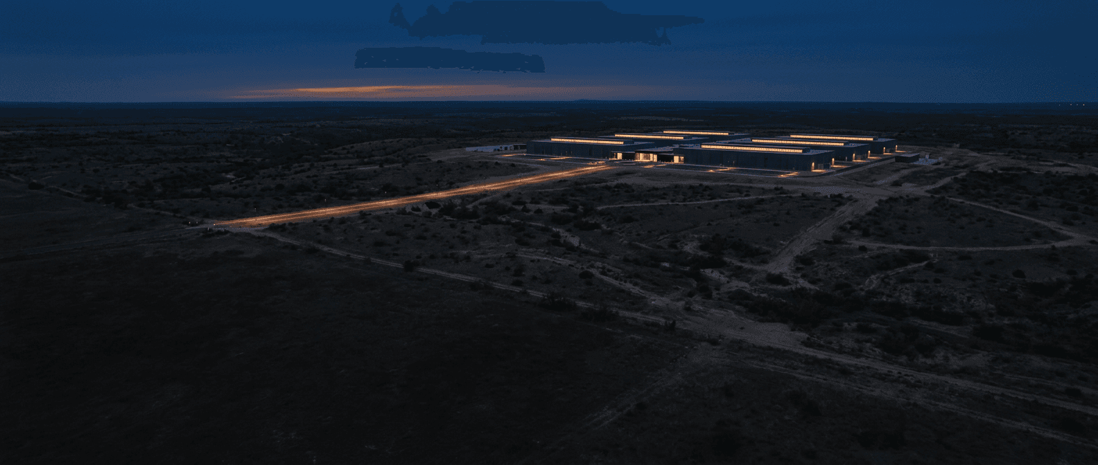 A single data center campus at blue hour seen across dark ranchland, one lit access road threading toward it
